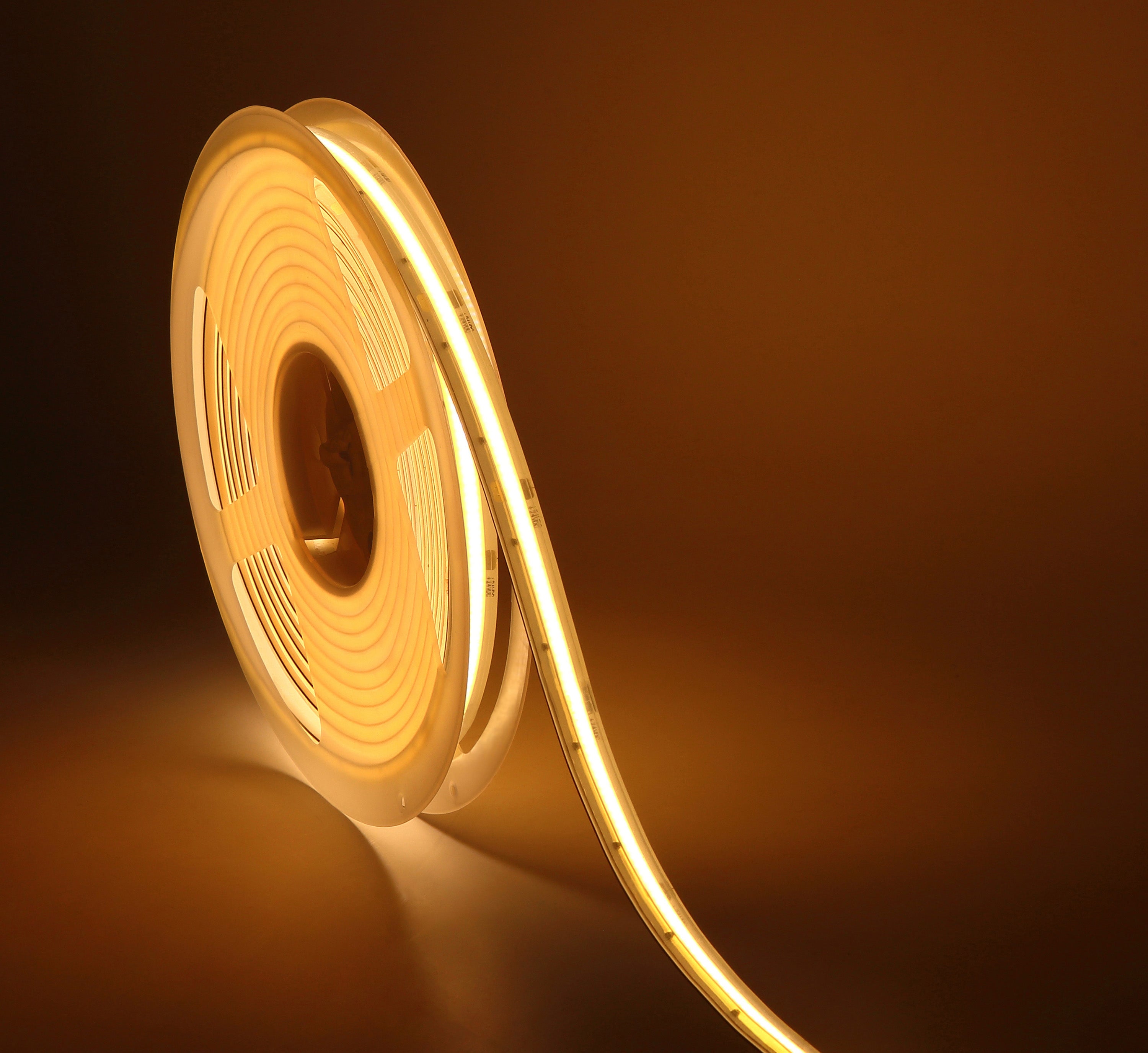 A square-shaped image of a single strip of LED tape light curled loosely in the center against a dark background, emitting a warm golden glow that reflects off the surface beneath it.