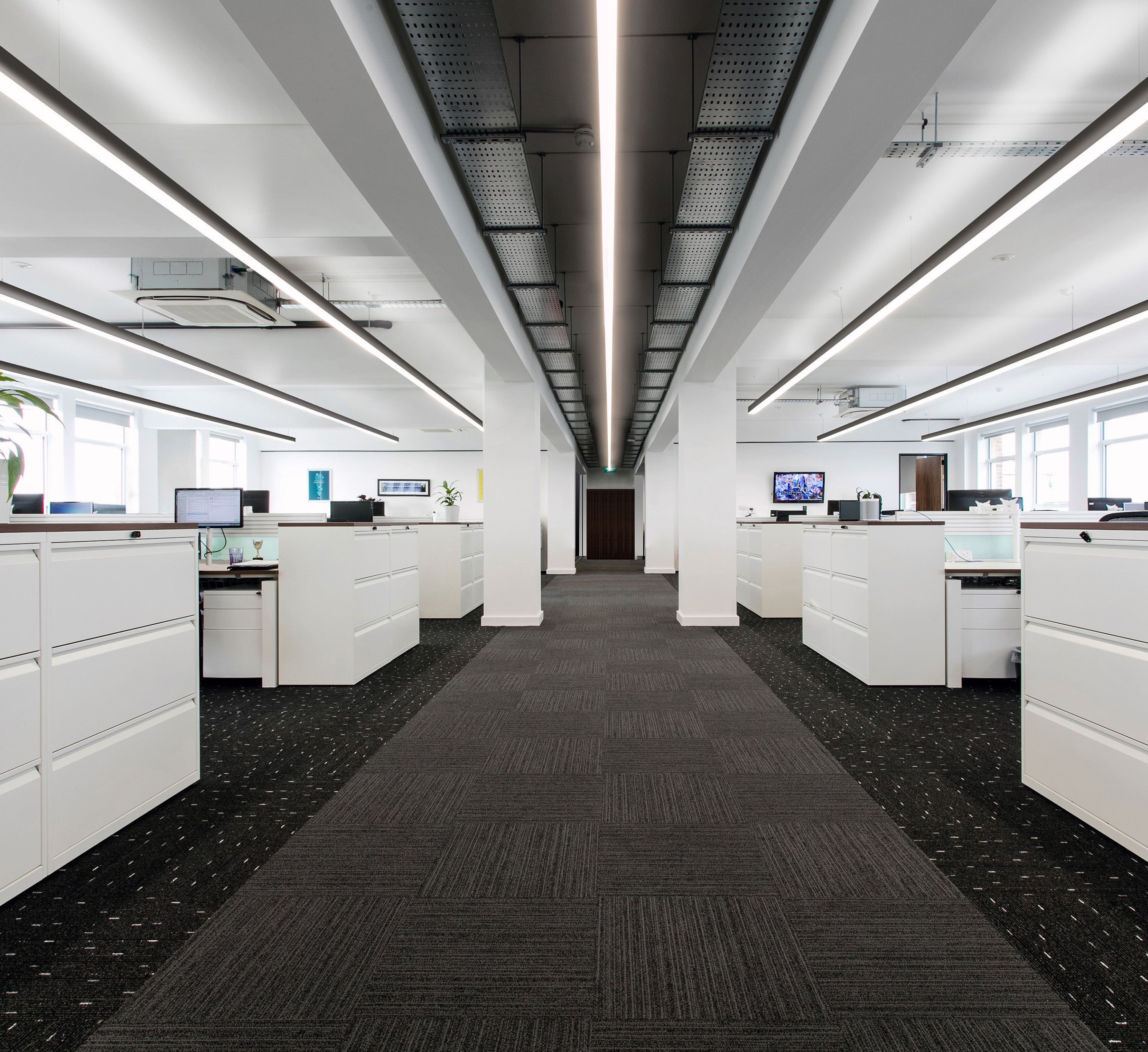 Spacious and brightly lit modern office interior with linear LED lights embedded in the ceiling, reflecting on the glossy white desks below. The room is lined with large windows, and green potted plants add a touch of nature to the workplace. The office design combines a minimalist aesthetic with a functional layout, featuring ergonomic chairs and organized workstations.
