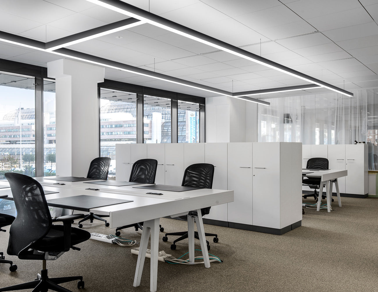 Extended and suspended linear light fixtures formed by GL LED L8070 T-shaped and 4ft straight linear light fixtures illuminate an office area.