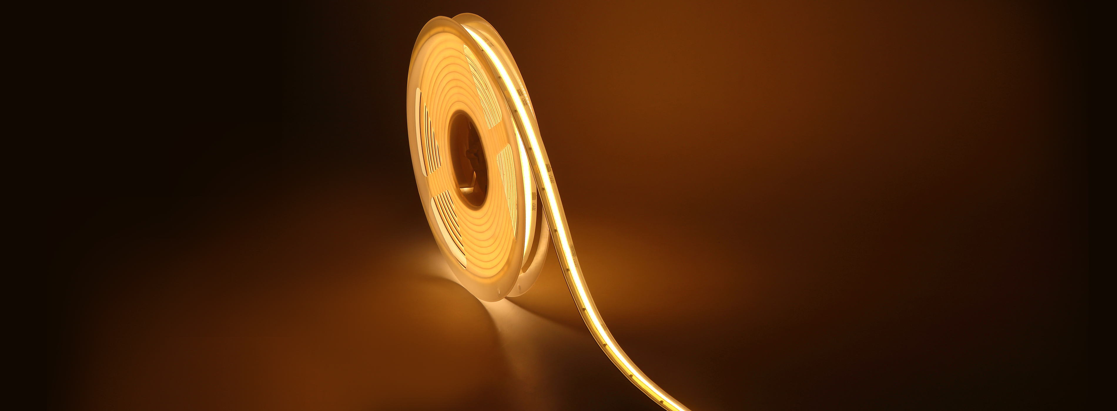 A wide panoramic image of a single strip of LED tape light curled loosely in the center against a dark background, emitting a warm golden glow that reflects off the surface beneath it.
