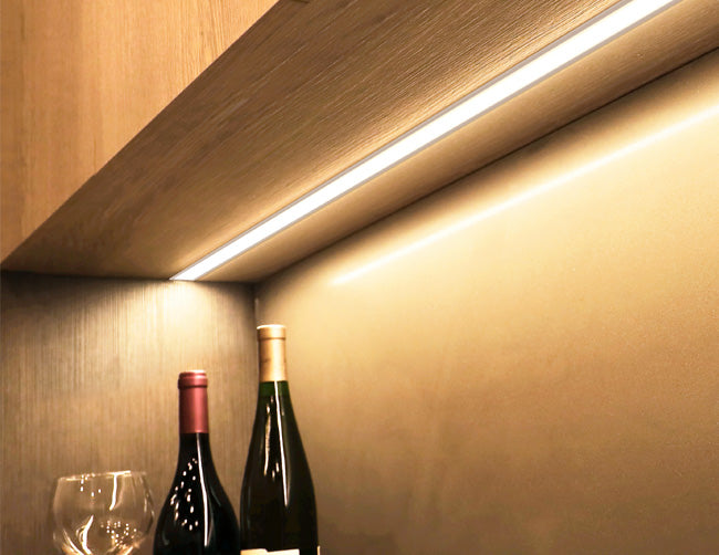 An LED strip light fixture is installed under a cabinet and illuminating the space. Two bottles of wine and a wine glass are shown below the undercabinet light.