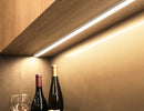 An LED strip light fixture is surface mounted under a cabinet and illuminating the space. Two bottles of wine and a wine glass are shown below the undercabinet light.