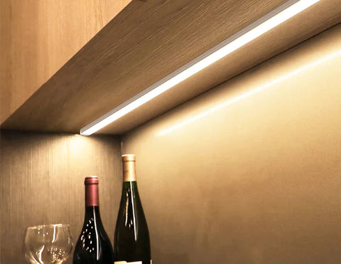 An LED strip light fixture is surface mounted under a cabinet and illuminating the space. Two bottles of wine and a wine glass are shown below the undercabinet light.