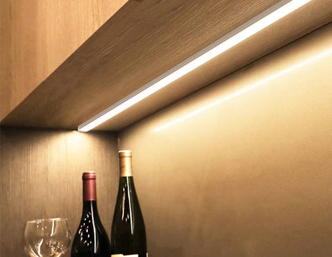 An LED strip light fixture is installed under a cabinet and illuminating the space. Two bottles of wine and a wine glass are shown below the undercabinet light.